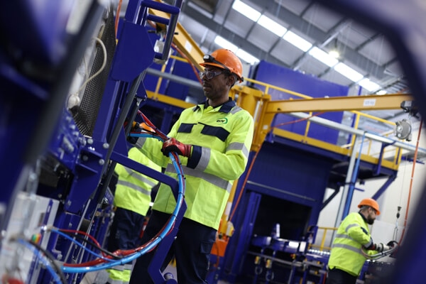 Two workers in a manufacturing or industrial setting. The foreground worker is wearing a high-visibility yellow and blue safety jacket, safety goggles, and an orange hard hat. He is handling cables connected to machinery.